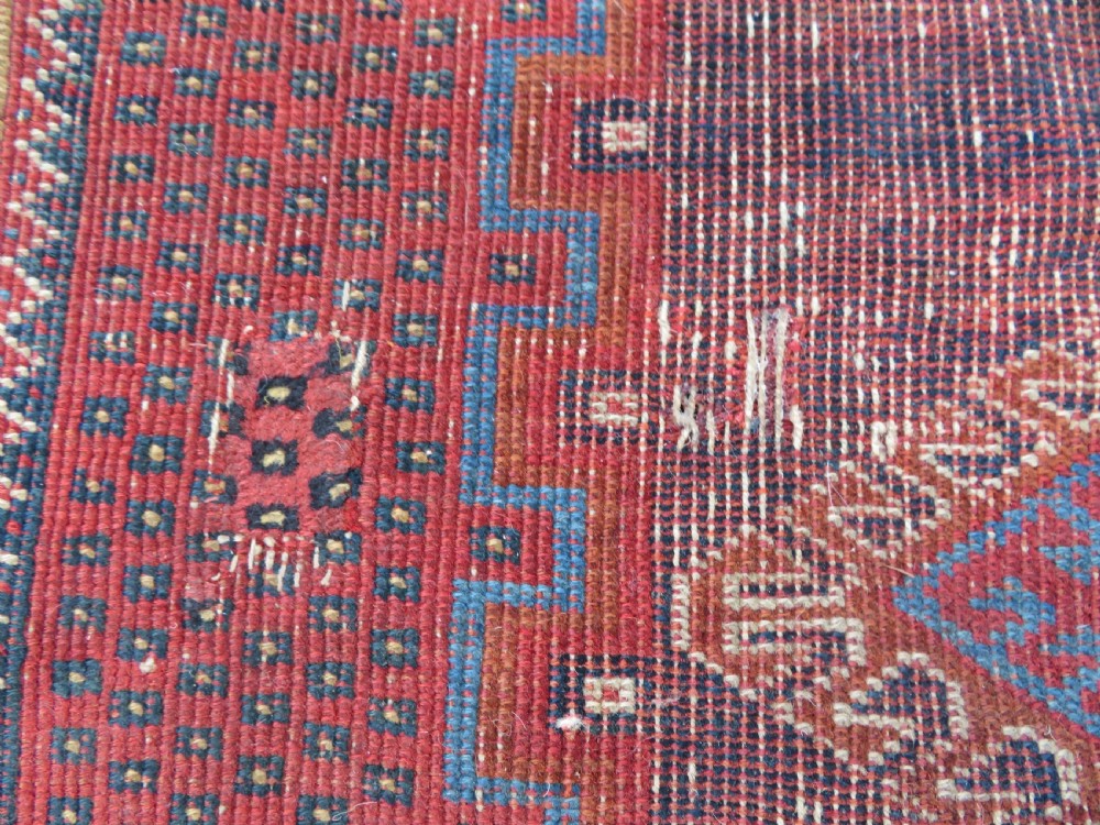 A rectangular hand-knotted wool rug featuring three diamond medallions on a dark central field surrounded by a patterned terracotta red background and geometric borders. View V