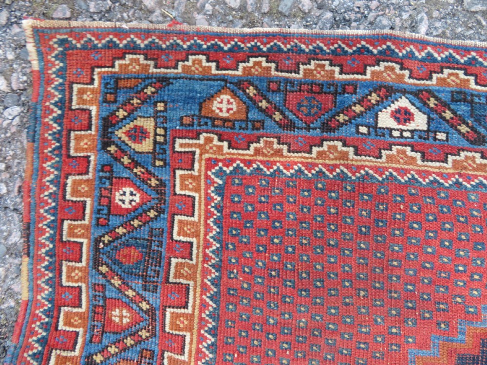 A rectangular hand-knotted wool rug featuring three diamond medallions on a dark central field surrounded by a patterned terracotta red background and geometric borders. View VI