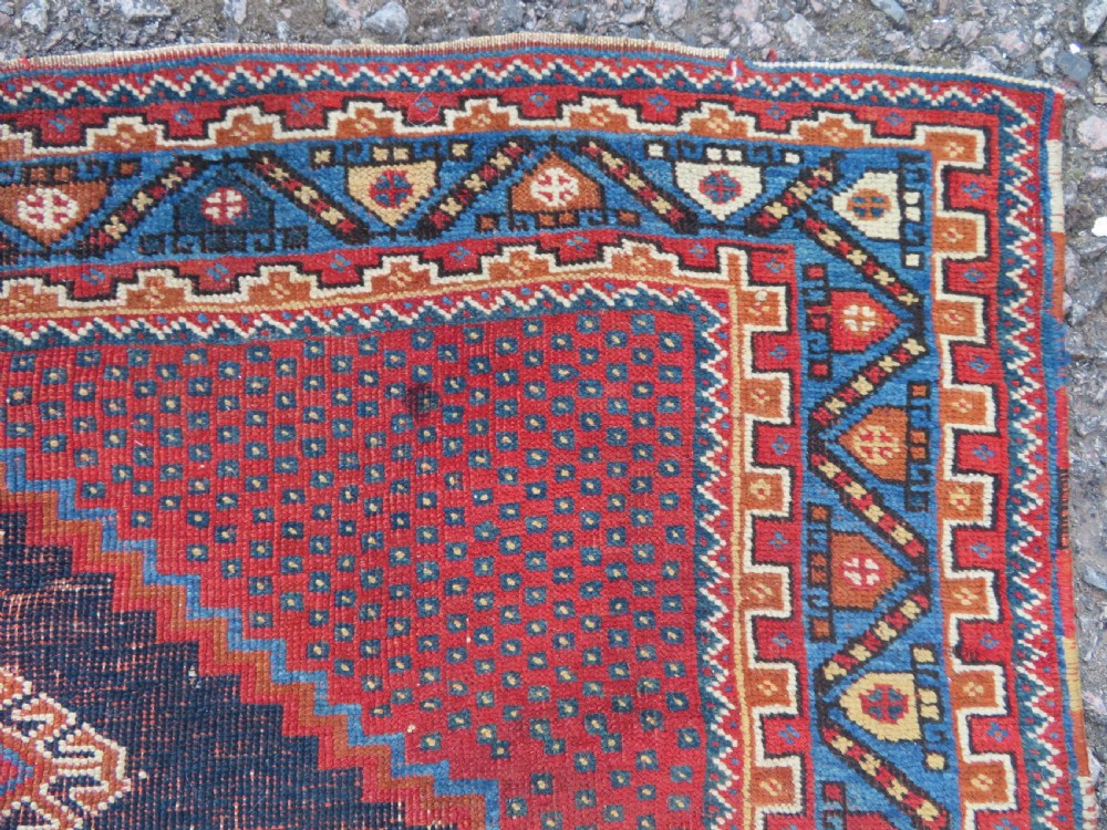 A rectangular hand-knotted wool rug featuring three diamond medallions on a dark central field surrounded by a patterned terracotta red background and geometric borders. View VII