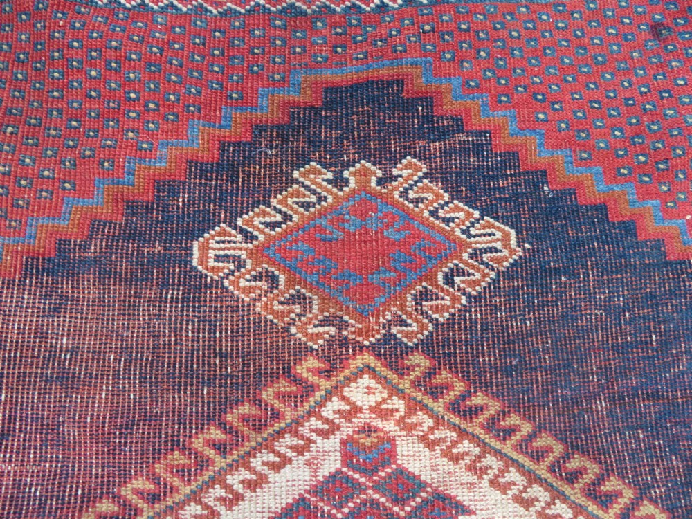 A rectangular hand-knotted wool rug featuring three diamond medallions on a dark central field surrounded by a patterned terracotta red background and geometric borders. View X