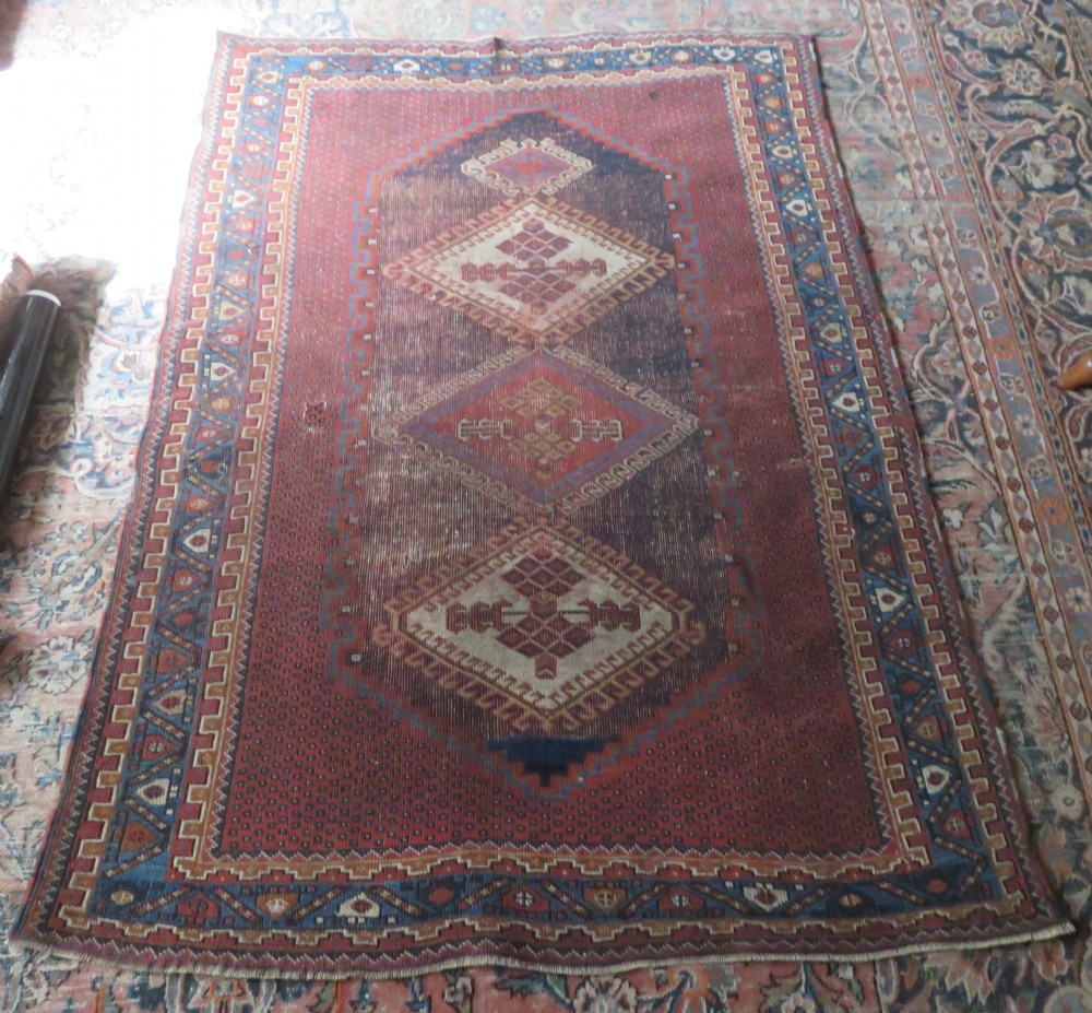 A rectangular hand-knotted wool rug featuring three diamond medallions on a dark central field surrounded by a patterned terracotta red background and geometric borders. View XV