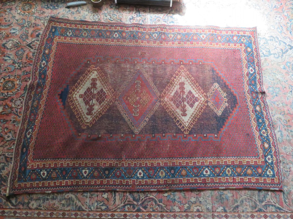 A rectangular hand-knotted wool rug featuring three diamond medallions on a dark central field surrounded by a patterned terracotta red background and geometric borders. View XVI