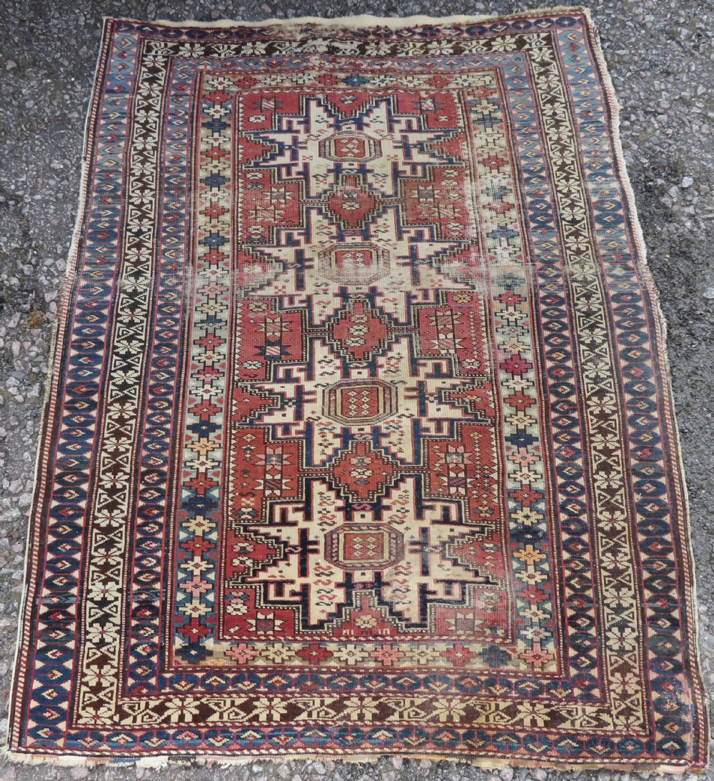A distressed antique hand-knotted Caucasian Daghestan wool rug featuring geometric medallions and intricate borders in terracotta red, cream, and indigo blue.