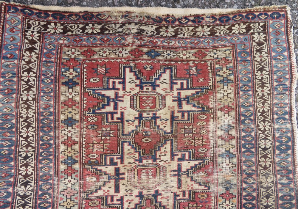 A distressed antique hand-knotted Caucasian Daghestan wool rug featuring geometric medallions and intricate borders in terracotta red, cream, and indigo blue. View II