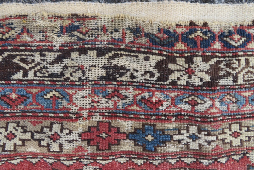 A distressed antique hand-knotted Caucasian Daghestan wool rug featuring geometric medallions and intricate borders in terracotta red, cream, and indigo blue. View III