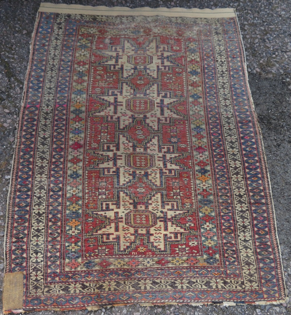 A distressed antique hand-knotted Caucasian Daghestan wool rug featuring geometric medallions and intricate borders in terracotta red, cream, and indigo blue. View IX