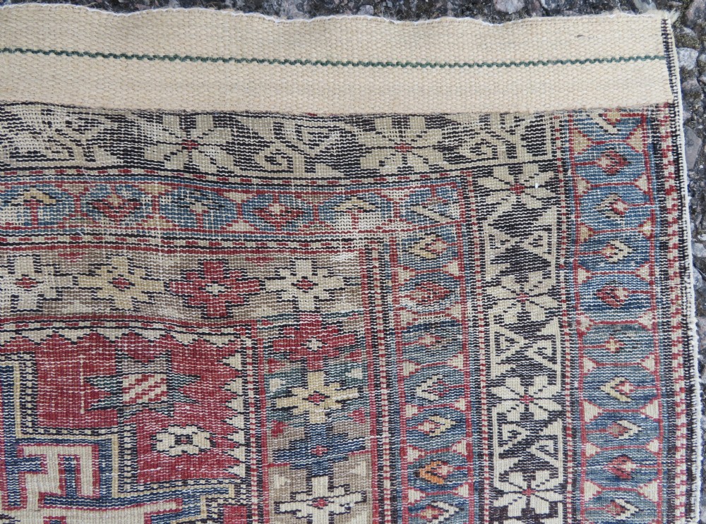 A distressed antique hand-knotted Caucasian Daghestan wool rug featuring geometric medallions and intricate borders in terracotta red, cream, and indigo blue. View X