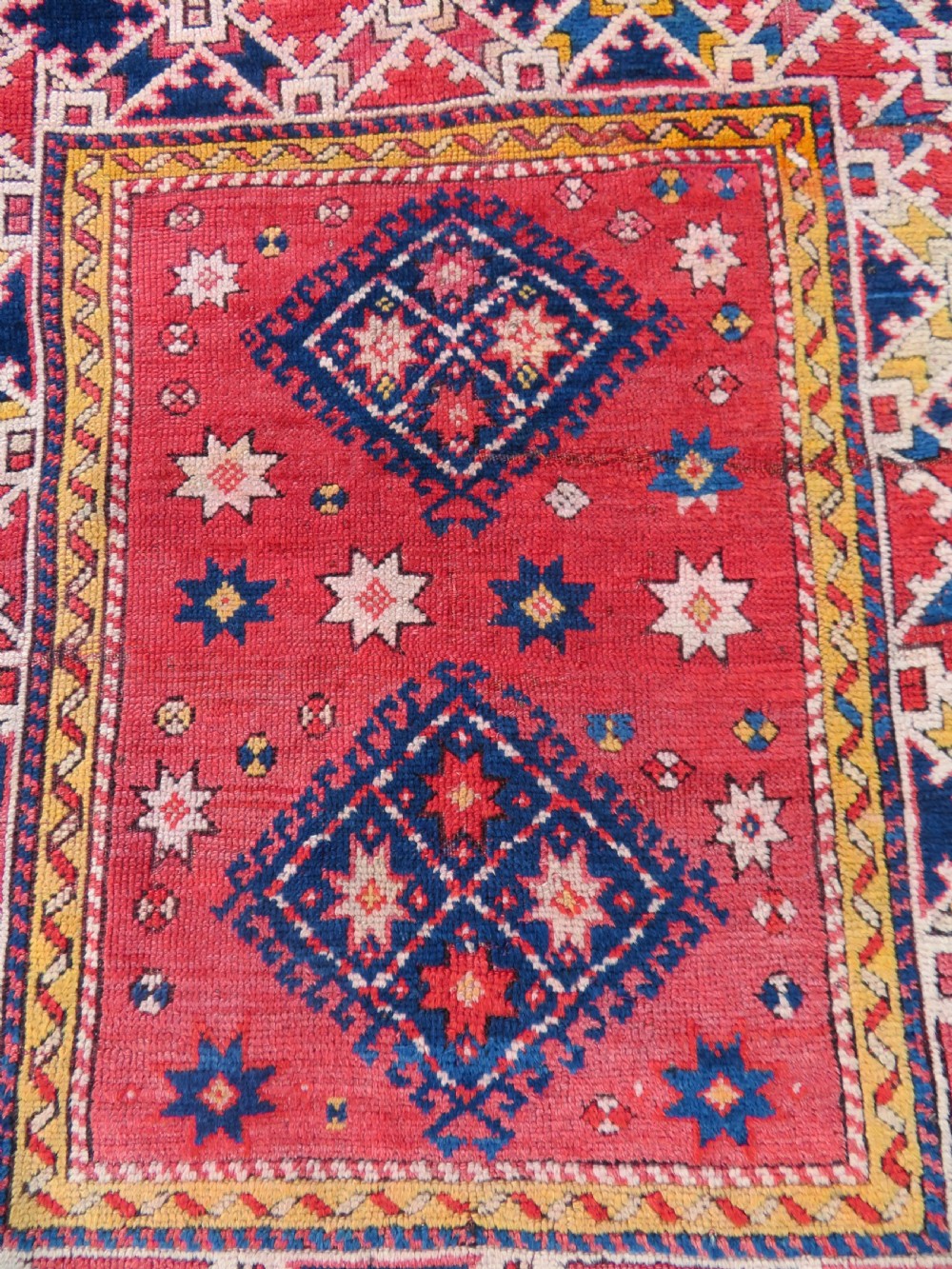 An antique hand-knotted wool rug featuring geometric diamond medallions in red, dark blue, yellow, and cream with a detailed border pattern. View II