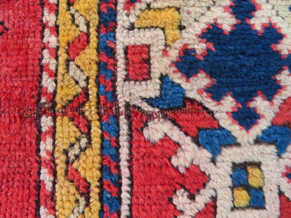 An antique hand-knotted wool rug featuring geometric diamond medallions in red, dark blue, yellow, and cream with a detailed border pattern. View VI
