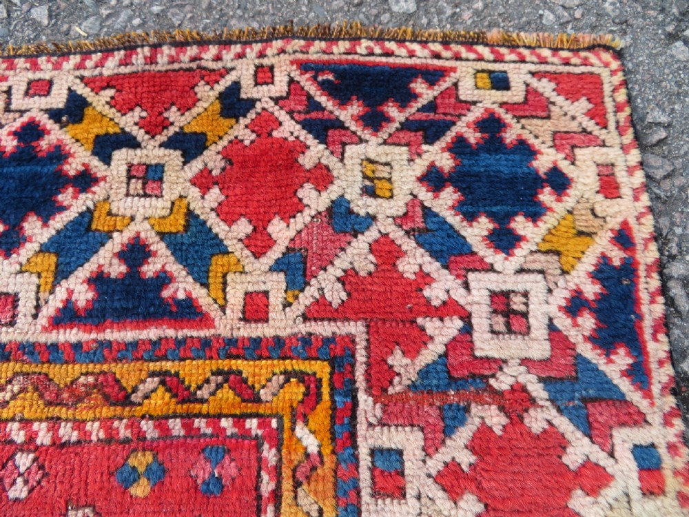 An antique hand-knotted wool rug featuring geometric diamond medallions in red, dark blue, yellow, and cream with a detailed border pattern. View VII