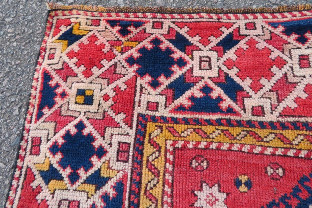 An antique hand-knotted wool rug featuring geometric diamond medallions in red, dark blue, yellow, and cream with a detailed border pattern. View VIII