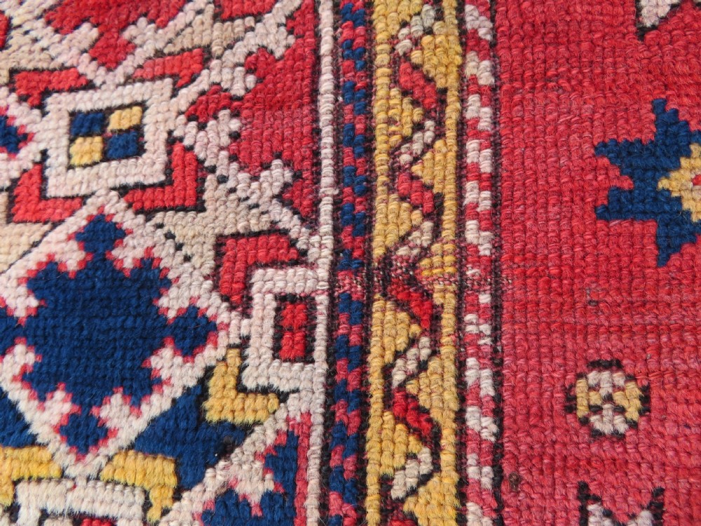 An antique hand-knotted wool rug featuring geometric diamond medallions in red, dark blue, yellow, and cream with a detailed border pattern. View X