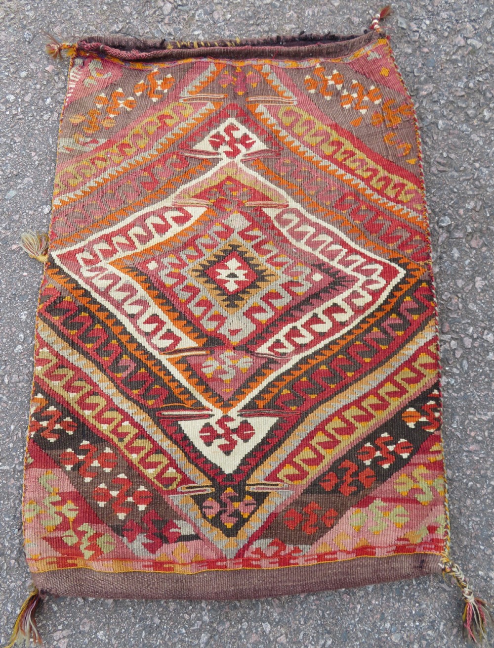 A flatwoven Anatolian kilim grain sack featuring geometric diamond motifs in red, orange, cream, and brown wool with corner tassels.
