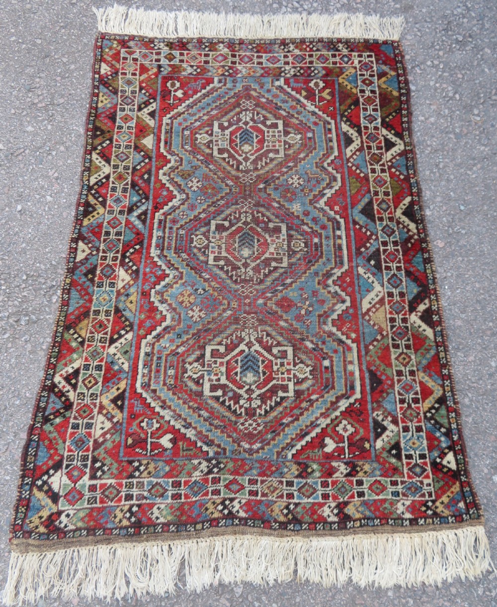 A small hand-knotted wool rug featuring three diamond geometric medallions on a blue and red field surrounded by detailed geometric borders with fringe at the ends.