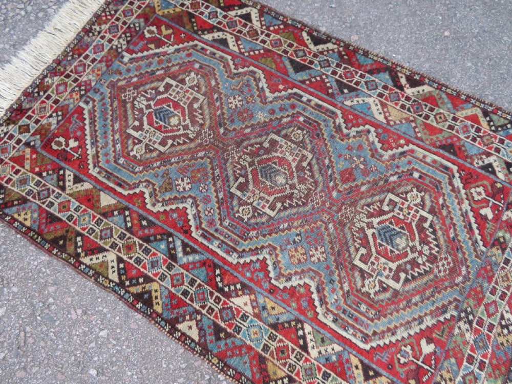 A small hand-knotted wool rug featuring three diamond geometric medallions on a blue and red field surrounded by detailed geometric borders with fringe at the ends. View II