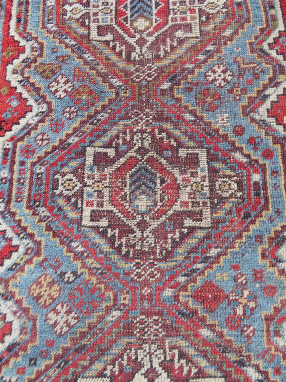 A small hand-knotted wool rug featuring three diamond geometric medallions on a blue and red field surrounded by detailed geometric borders with fringe at the ends. View IV