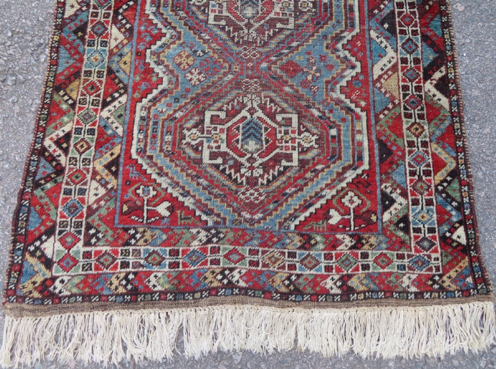 A small hand-knotted wool rug featuring three diamond geometric medallions on a blue and red field surrounded by detailed geometric borders with fringe at the ends. View V