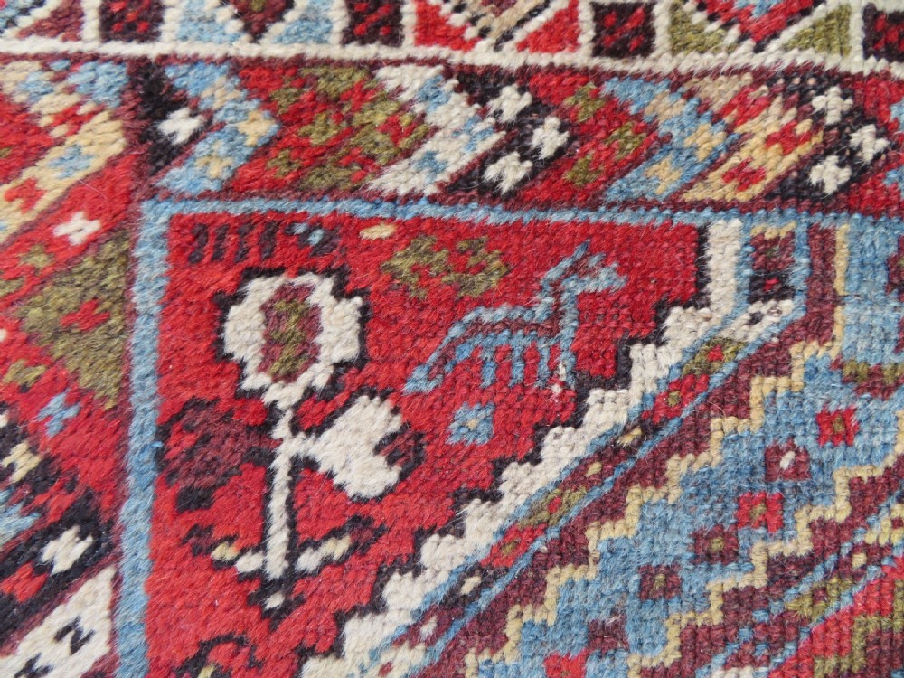 A small hand-knotted wool rug featuring three diamond geometric medallions on a blue and red field surrounded by detailed geometric borders with fringe at the ends. View VI