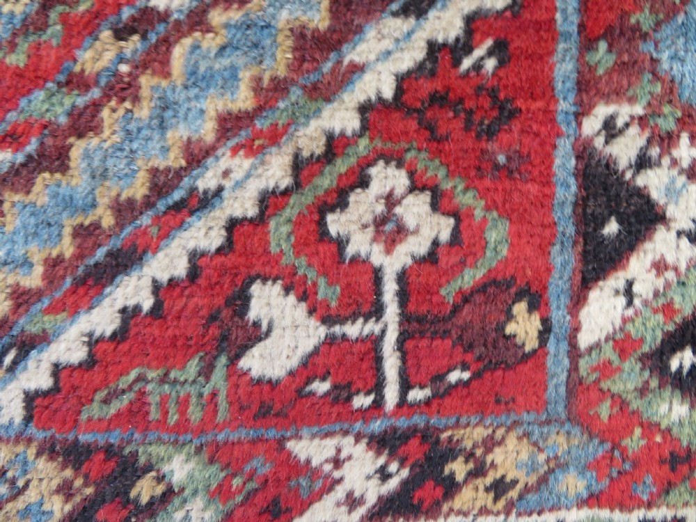 A small hand-knotted wool rug featuring three diamond geometric medallions on a blue and red field surrounded by detailed geometric borders with fringe at the ends. View VII