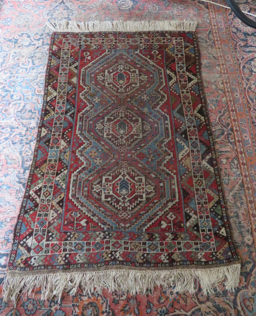 A small hand-knotted wool rug featuring three diamond geometric medallions on a blue and red field surrounded by detailed geometric borders with fringe at the ends. View XVII