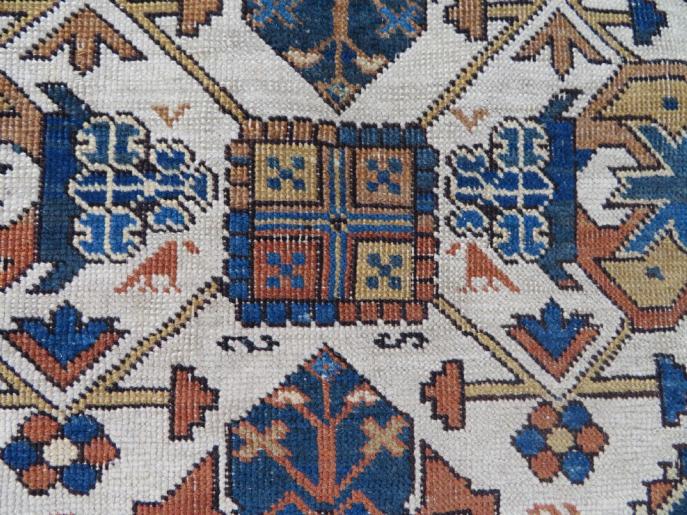 An antique hand-knotted Caucasian Kuba Seichur wool rug featuring a geometric white ground field surrounded by multi-layered patterned borders in blue, terracotta, and ochre. View V