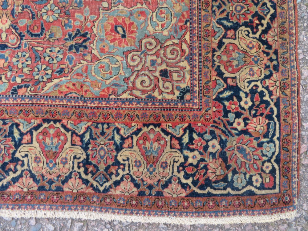 A rectangular hand-knotted wool Kashan rug featuring a central starburst medallion surrounded by dense floral patterns on a terracotta field with a navy border. View X