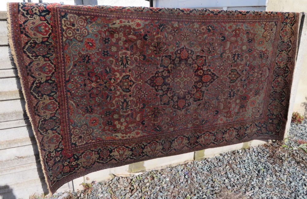 A rectangular hand-knotted wool Kashan rug featuring a central starburst medallion surrounded by dense floral patterns on a terracotta field with a navy border. View XX