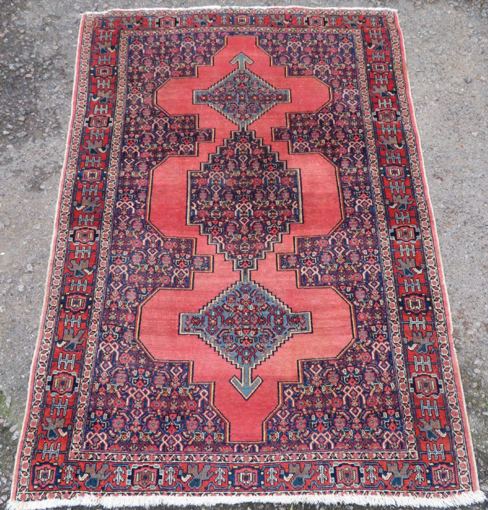 An antique West Persian Senneh wool rug featuring a navy blue stepped diamond medallion centered on a terracotta red field with intricate geometric borders.