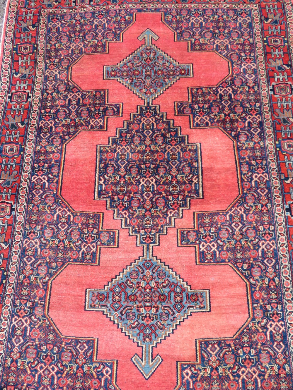 An antique West Persian Senneh wool rug featuring a navy blue stepped diamond medallion centered on a terracotta red field with intricate geometric borders. View III