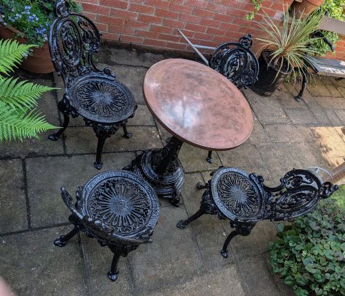 fig 1. A Victorian cast-iron garden set featuring a circular copper-topped pedestal table surrounded by four ornate black cast-iron chairs with foliage-patterned backrests.