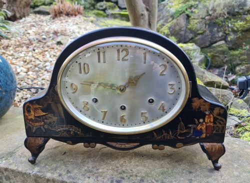 fig 1. An antique black lacquered mantel clock featuring hand-painted gilt chinoiserie scenes, an oval clock face with gold Arabic numerals, and brass bracket feet.