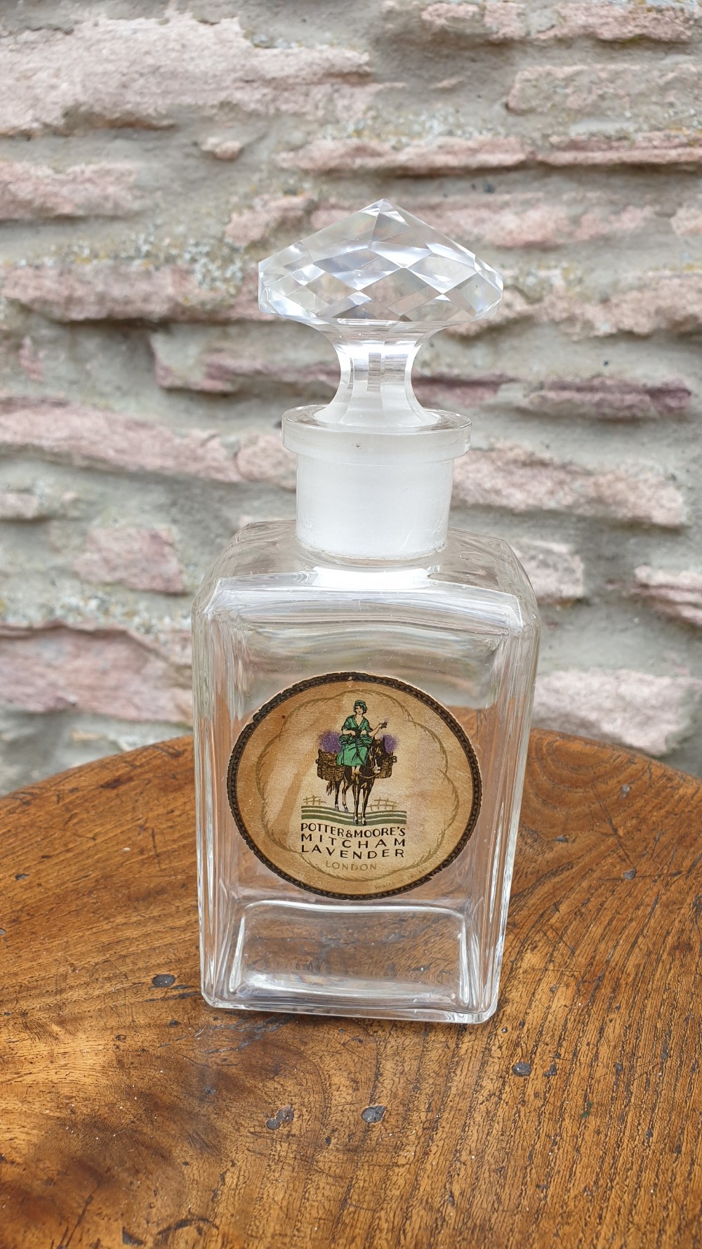 A clear glass rectangular perfume bottle with a faceted geometric stopper and an original circular paper label reading Potter and Moores Mitcham Lavender.