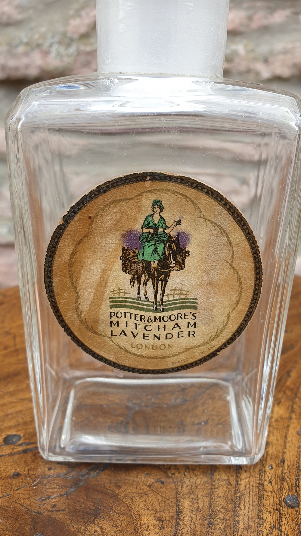 A clear glass rectangular perfume bottle with a faceted geometric stopper and an original circular paper label reading Potter and Moores Mitcham Lavender. View II
