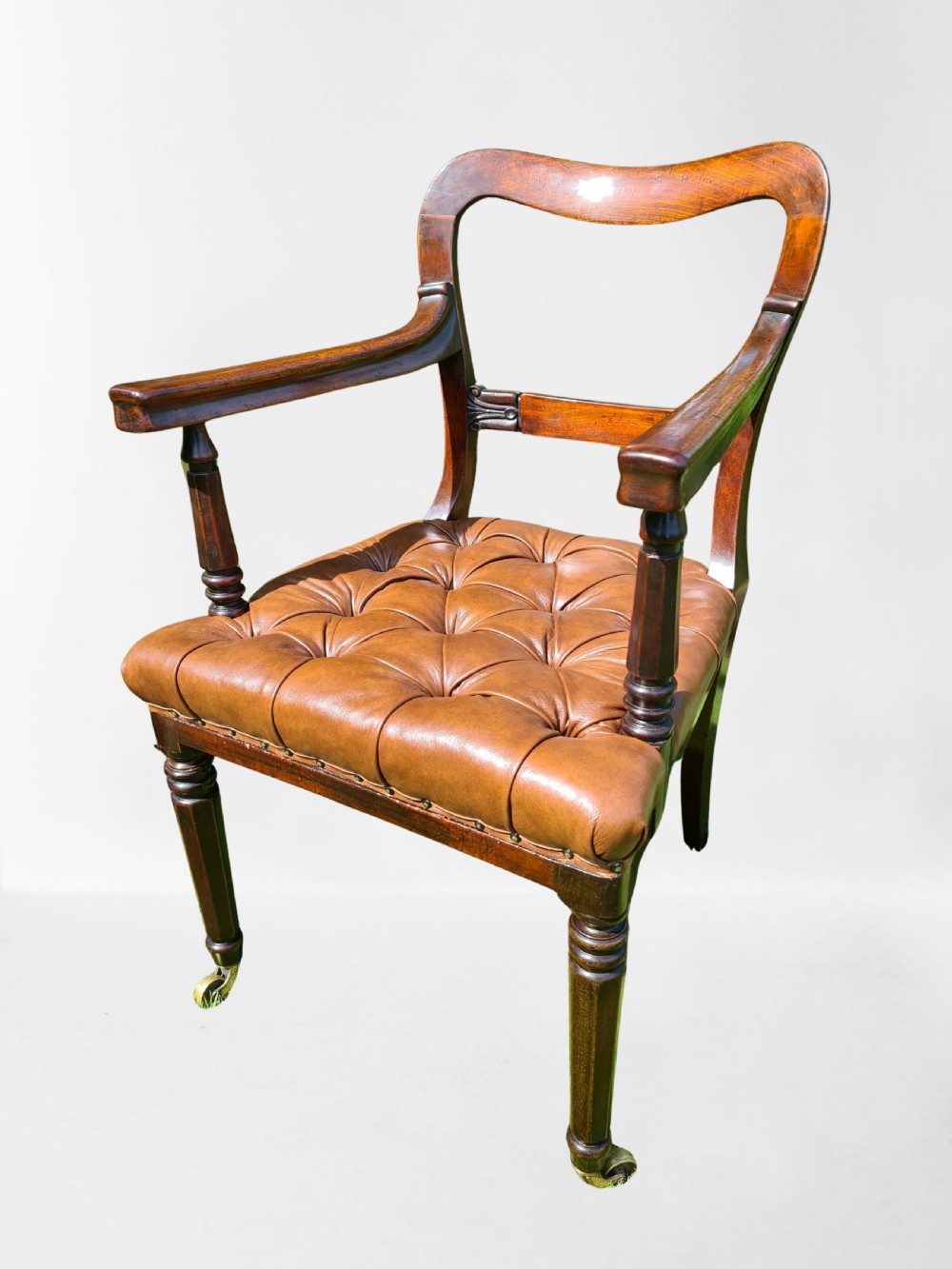 An early 19th-century mahogany desk armchair featuring a curved backrest, open arms, turned front legs on brass castors, and a button-tufted tan leather seat.