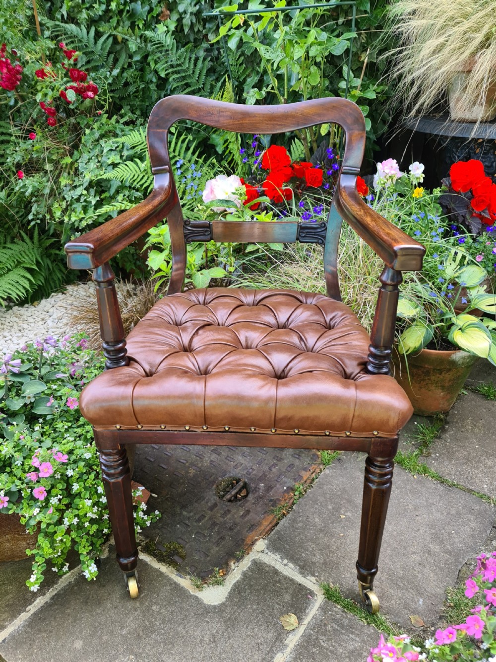 An early 19th-century mahogany desk armchair featuring a curved backrest, open arms, turned front legs on brass castors, and a button-tufted tan leather seat. View VII
