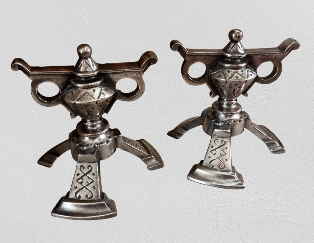 A pair of engraved steel fire dogs featuring urn-shaped tops, scroll side handles, geometric etchings, and tripod feet against a grey background.