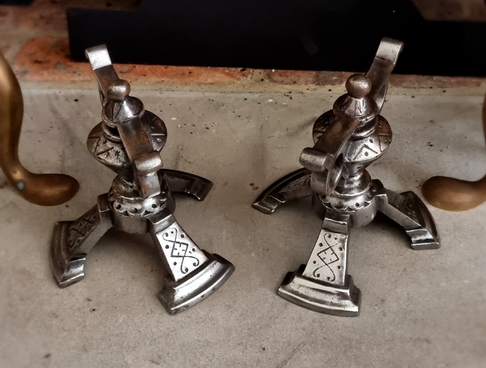 A pair of engraved steel fire dogs featuring urn-shaped tops, scroll side handles, geometric etchings, and tripod feet against a grey background. View III