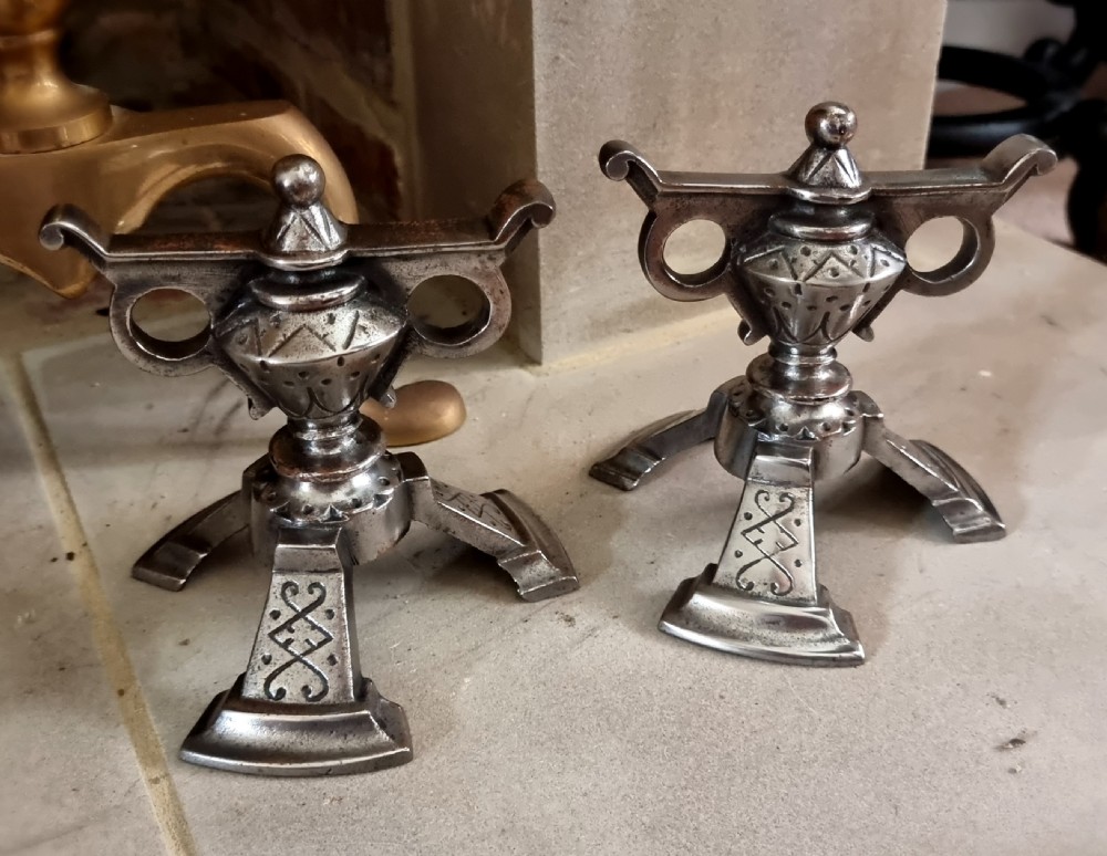 A pair of engraved steel fire dogs featuring urn-shaped tops, scroll side handles, geometric etchings, and tripod feet against a grey background. View IV