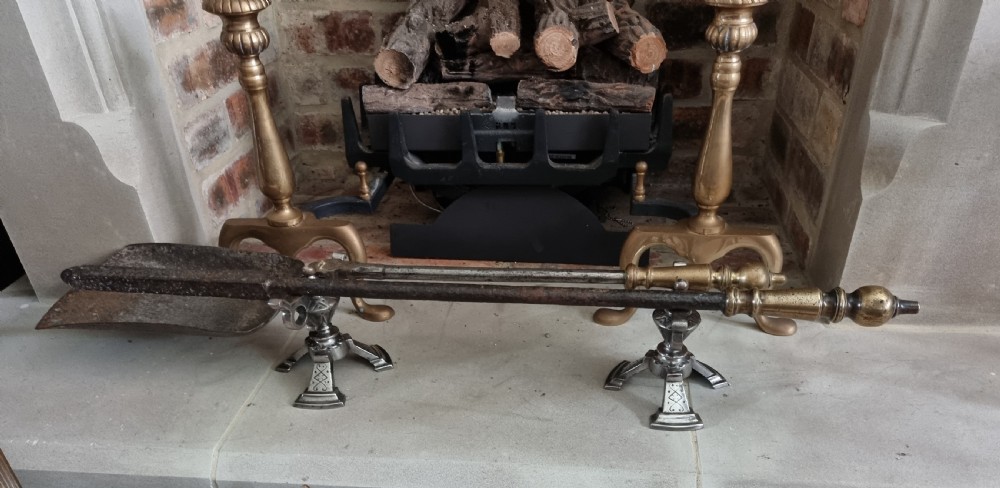 A pair of engraved steel fire dogs featuring urn-shaped tops, scroll side handles, geometric etchings, and tripod feet against a grey background. View V
