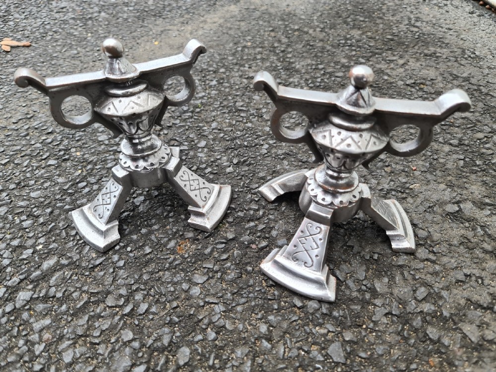 A pair of engraved steel fire dogs featuring urn-shaped tops, scroll side handles, geometric etchings, and tripod feet against a grey background. View VII