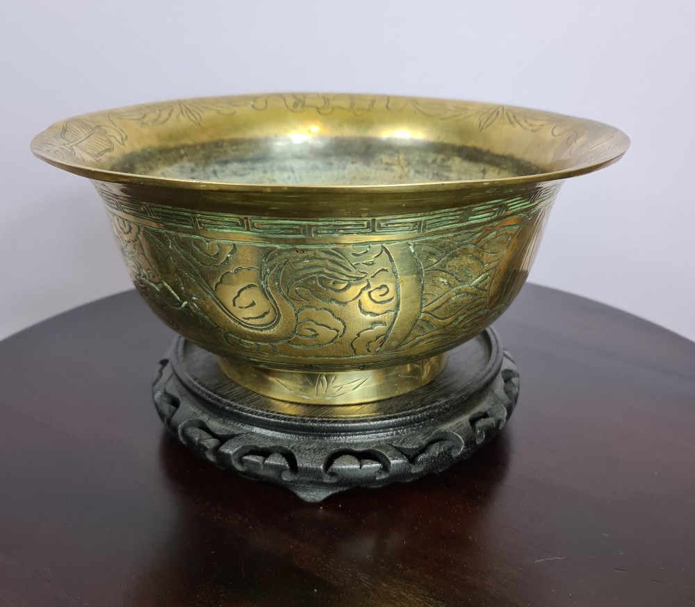 A large brass bowl featuring engraved dragon and cloud patterns with a flared rim, displayed on a dark carved wooden stand. View II