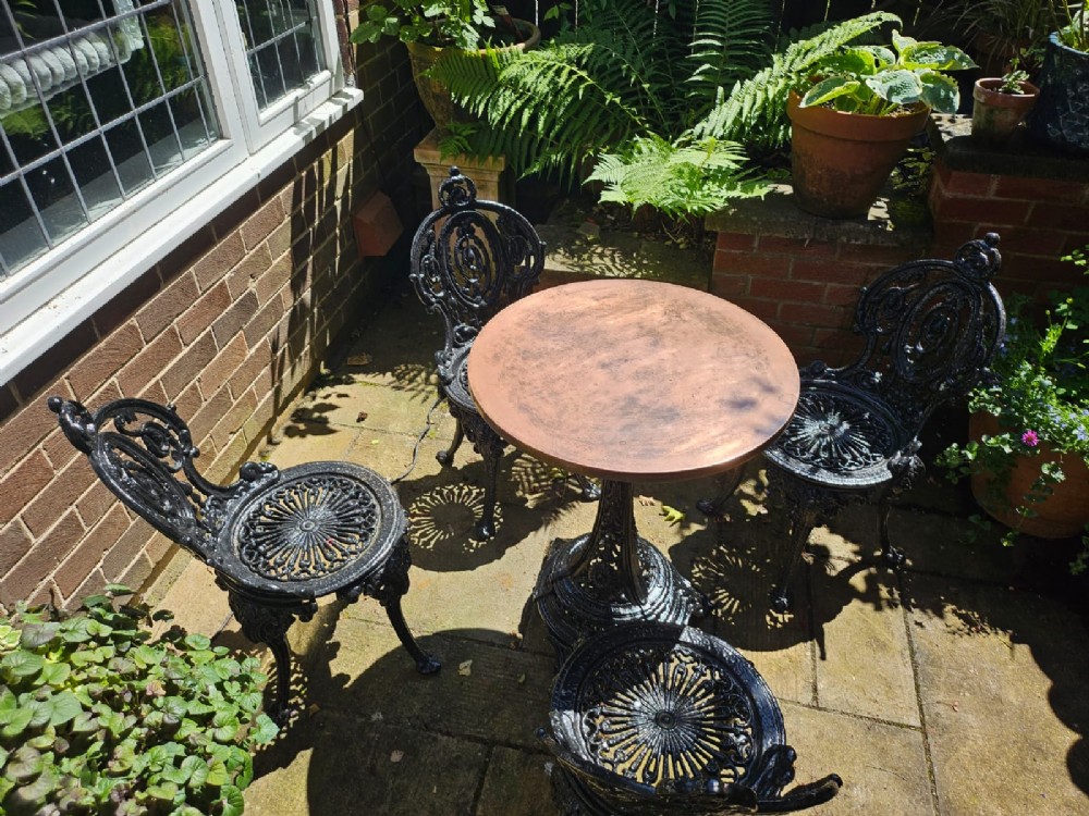 A Victorian cast iron garden set featuring a circular table with a copper top alongside four black ornate openwork chairs. View II