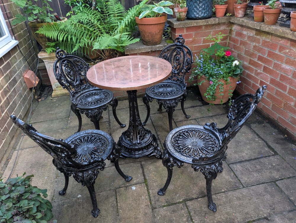 A Victorian cast iron garden set featuring a circular table with a copper top alongside four black ornate openwork chairs. View X