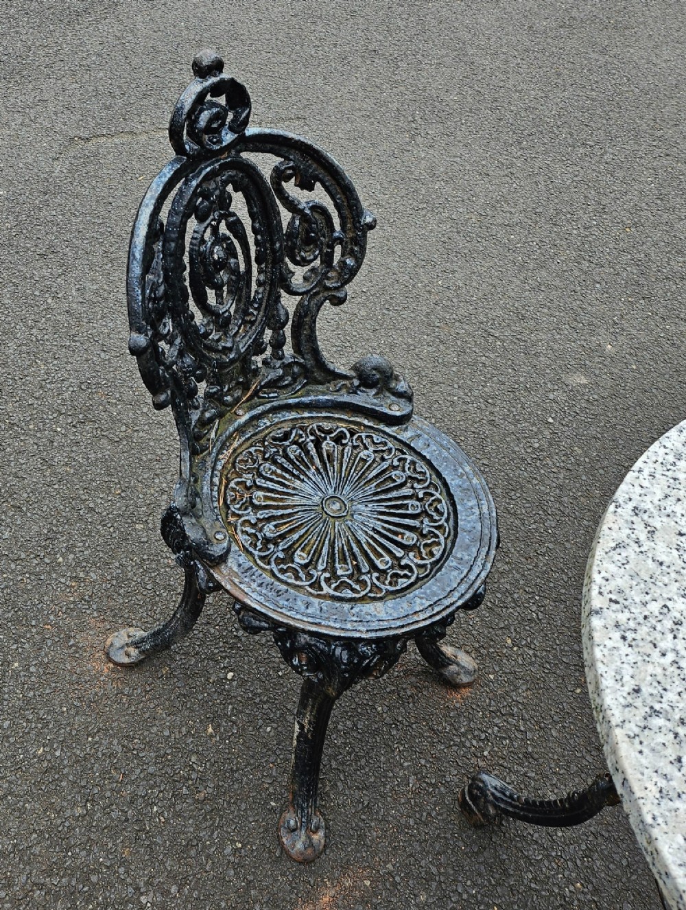 A black ornate cast-iron garden set featuring a circular stone-topped pedestal table and four matching pierced-back chairs. View V