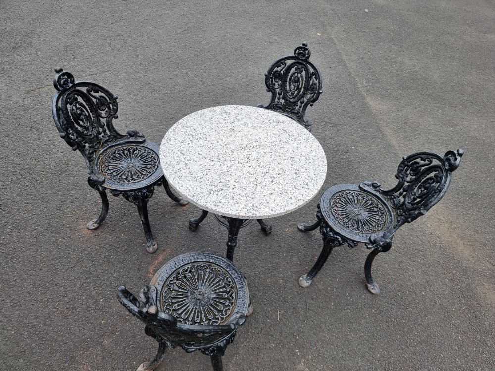 A black ornate cast-iron garden set featuring a circular stone-topped pedestal table and four matching pierced-back chairs. View IX
