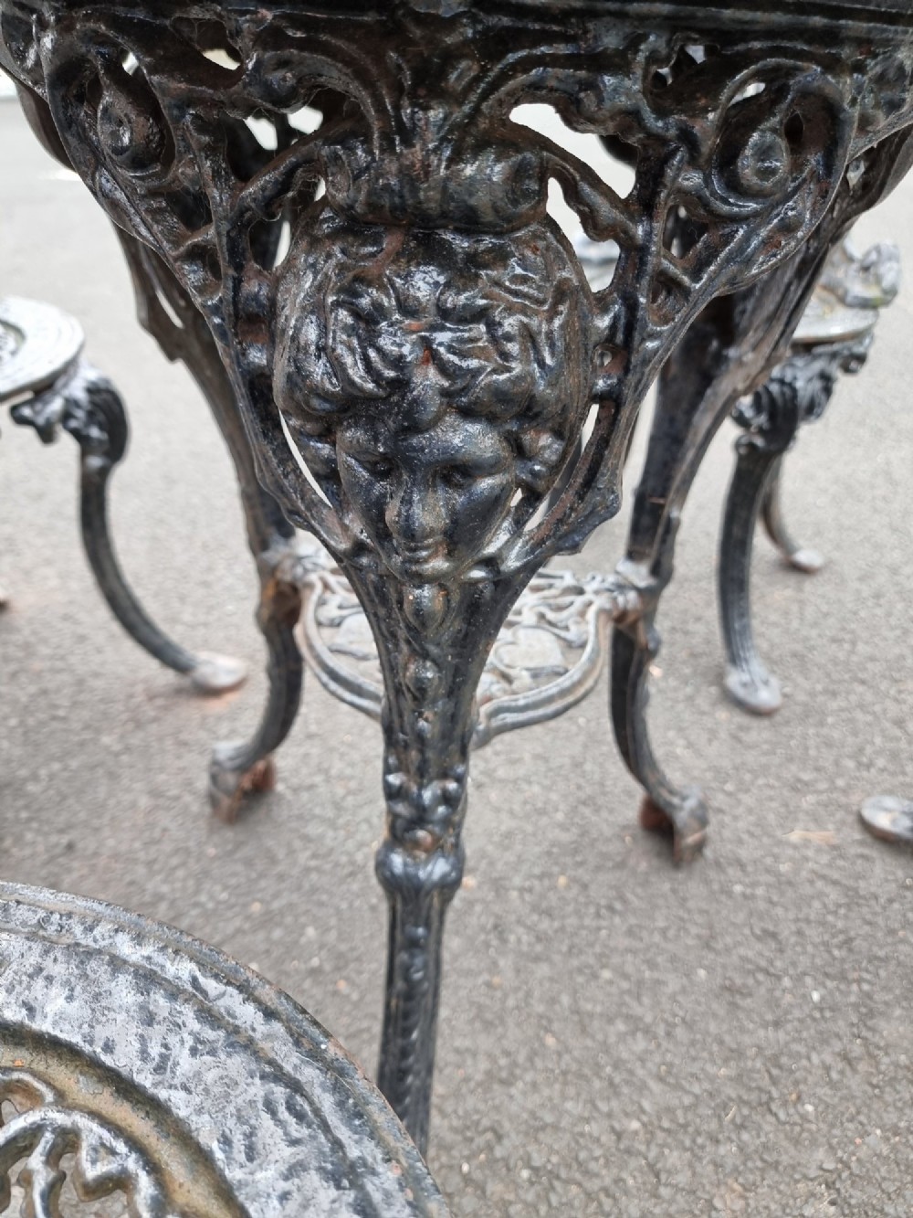 A black ornate cast-iron garden set featuring a circular stone-topped pedestal table and four matching pierced-back chairs. View X