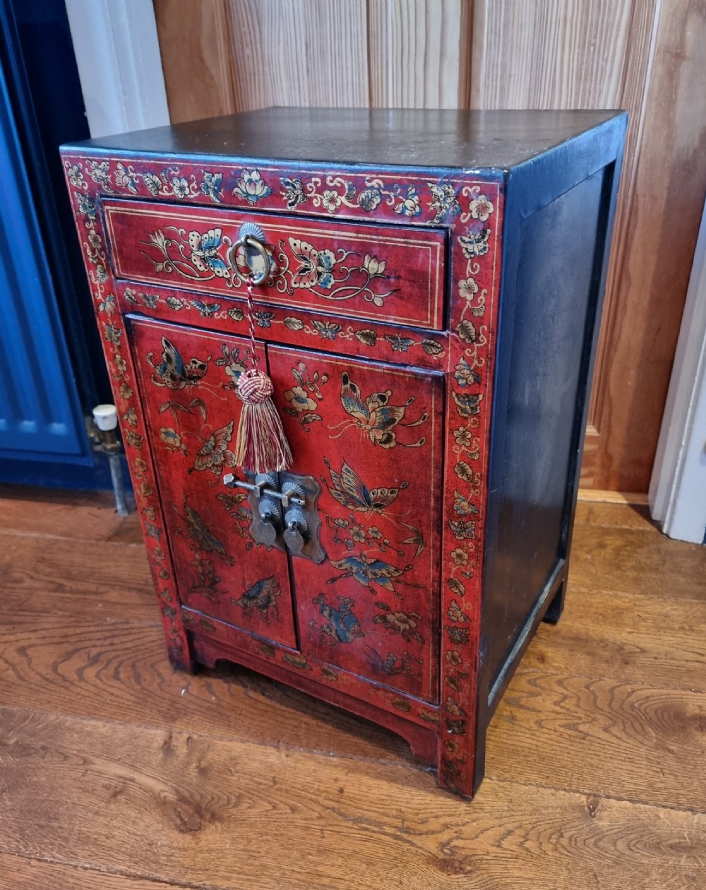 A small red and black hand-painted Chinoiserie cupboard featuring butterfly decoration, a single top drawer, two lower doors, and traditional brass hardware.
