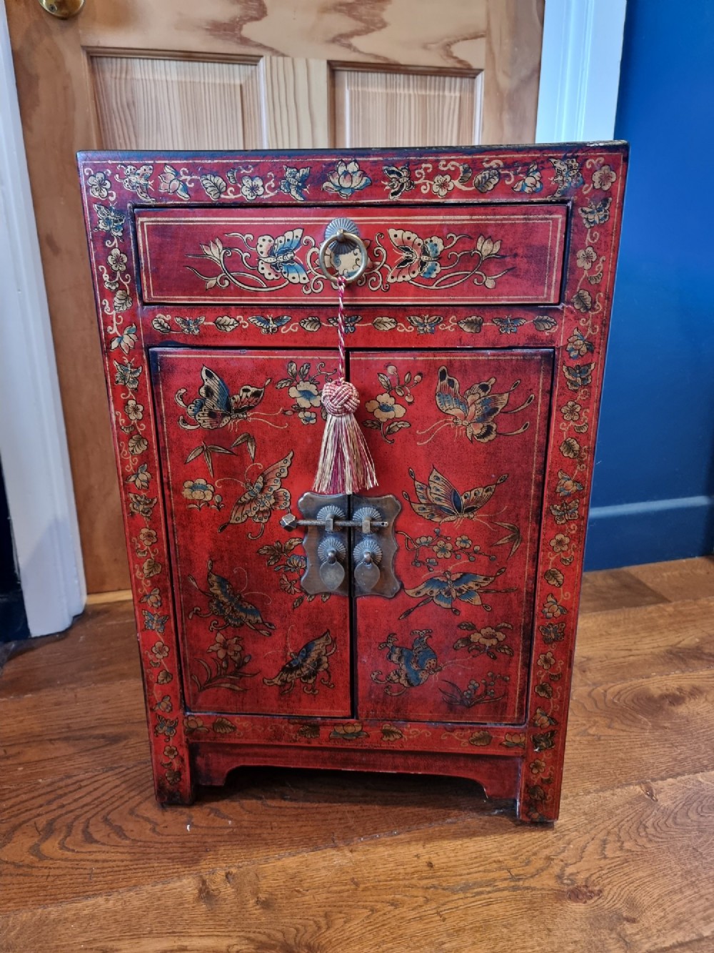 A small red and black hand-painted Chinoiserie cupboard featuring butterfly decoration, a single top drawer, two lower doors, and traditional brass hardware. View II