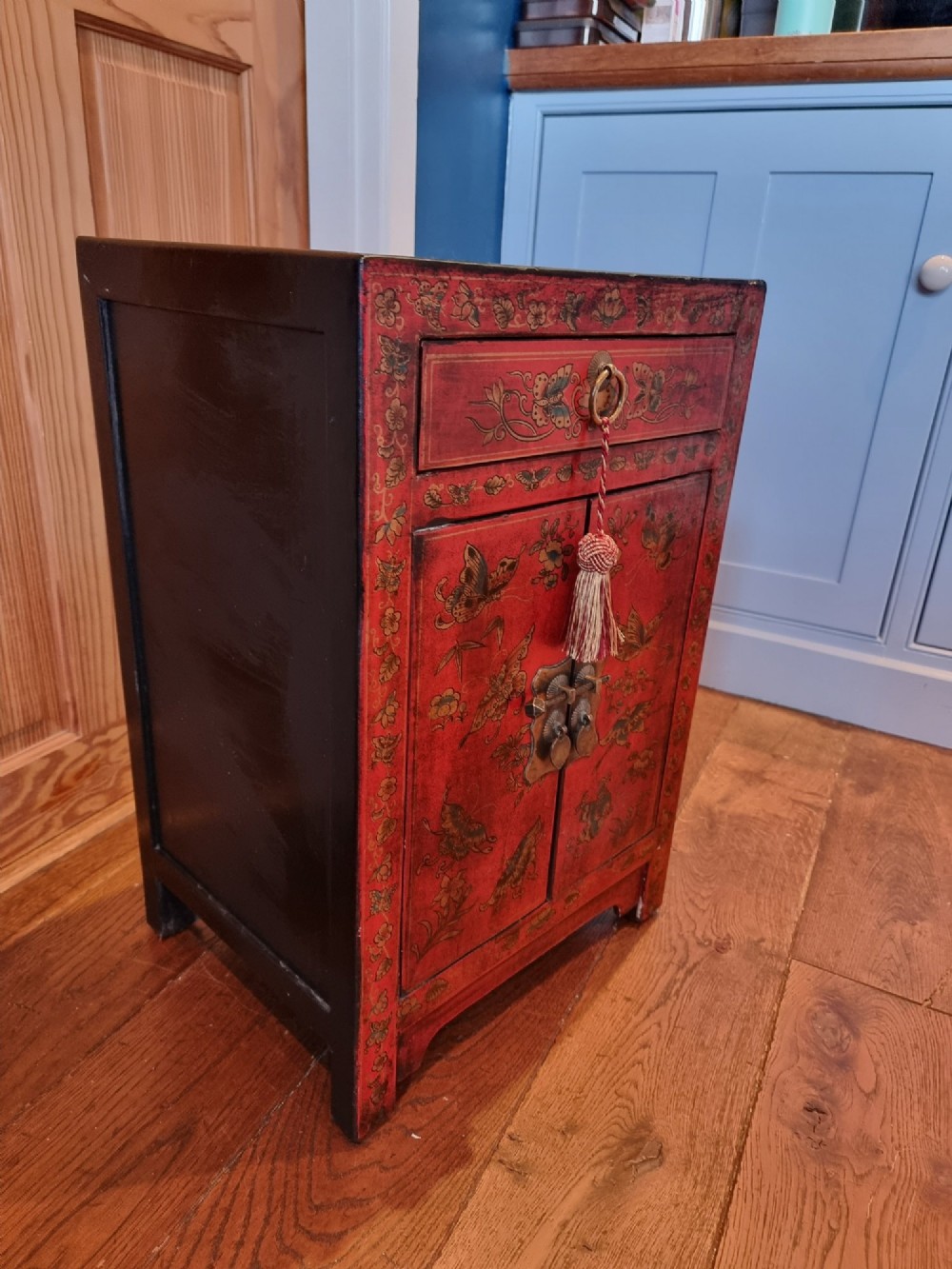 A small red and black hand-painted Chinoiserie cupboard featuring butterfly decoration, a single top drawer, two lower doors, and traditional brass hardware. View III
