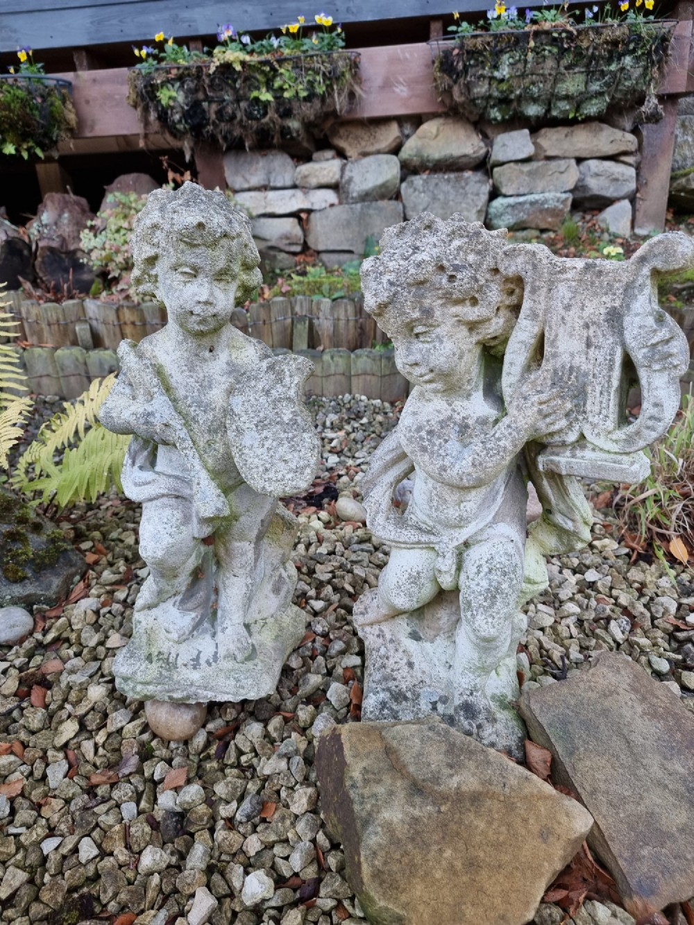 A pair of weathered grey reconstituted stone garden statues depicting classical musical cherubs holding instruments.